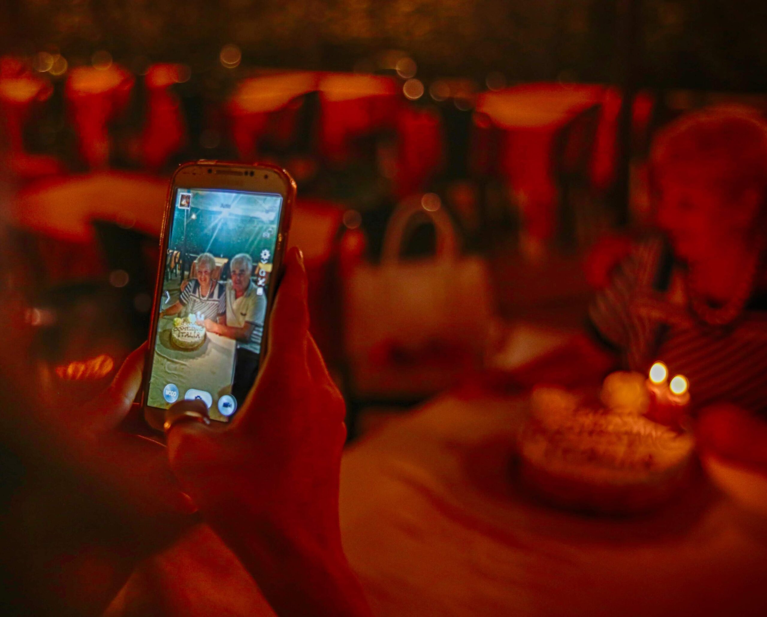 Mani che tengono uno smartphone che cattura l’immagine di due persone sorridenti accanto a una torta di compleanno con candeline accese.
